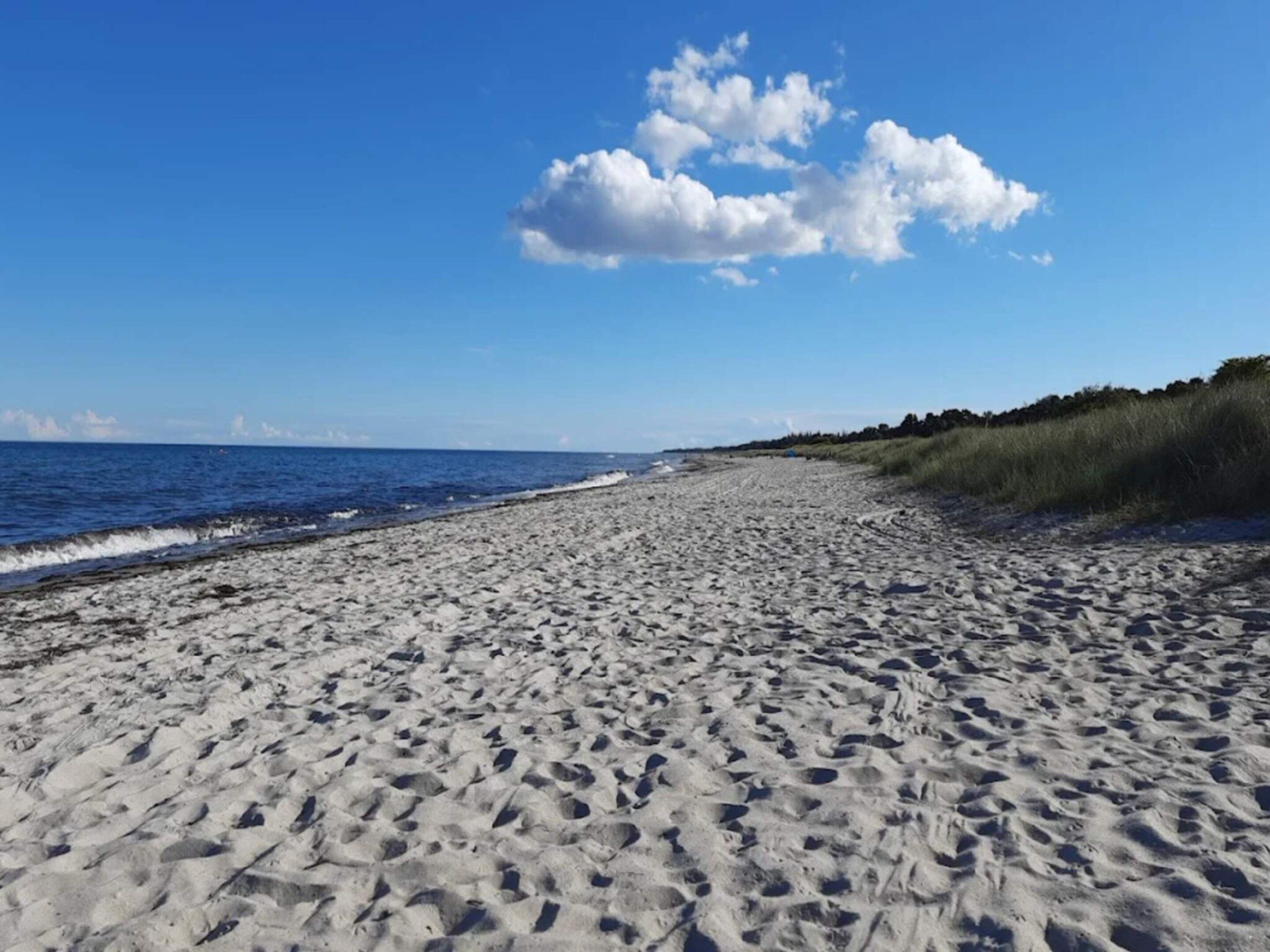 Een zandstrand met voetstappen die naar het water leiden, een heldere lucht en een grasveld aan de rechterkant.