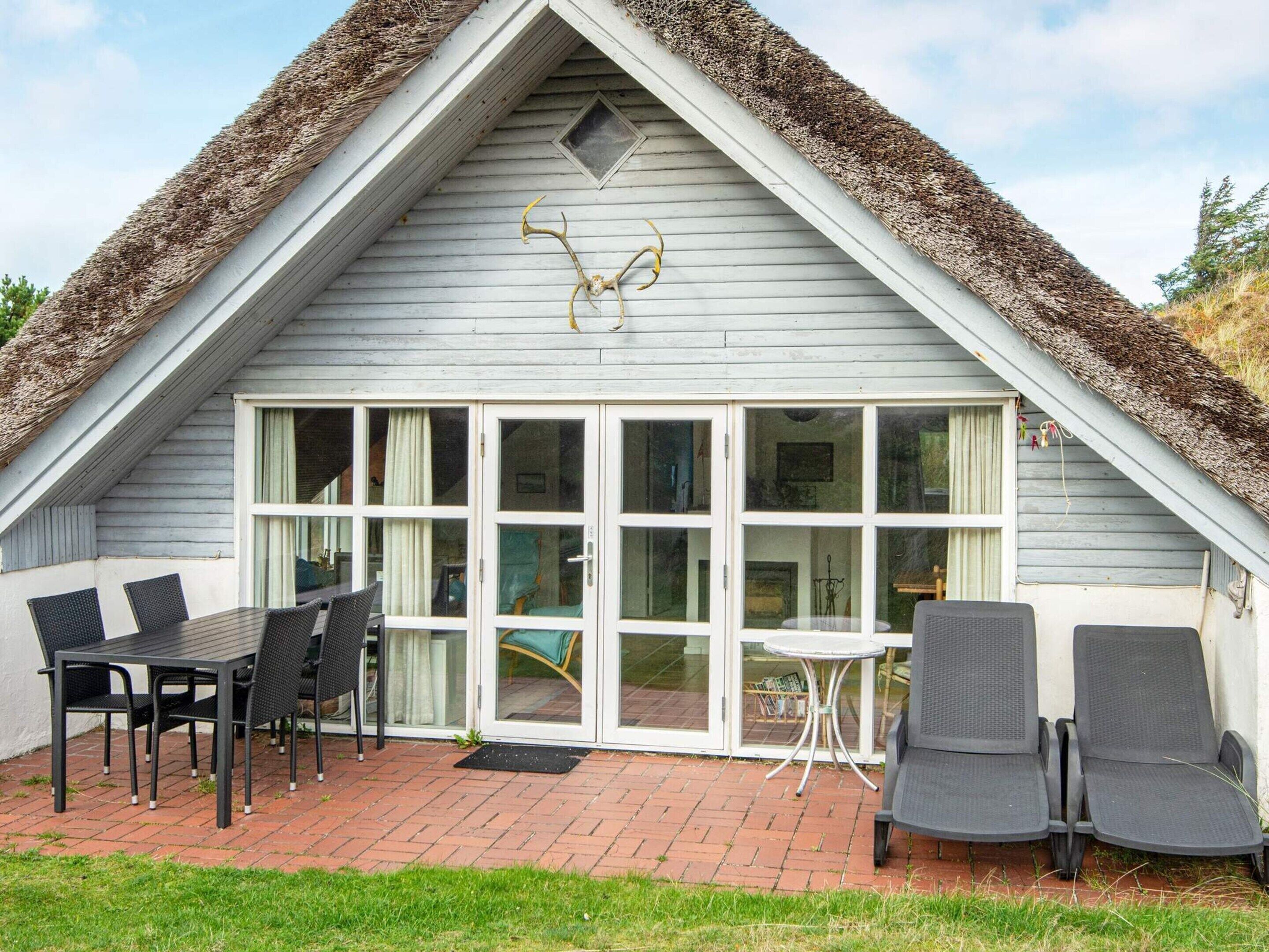 A thatched-roof cottage with a sliding glass door, a dining table with chairs, and a small round table with a chair.