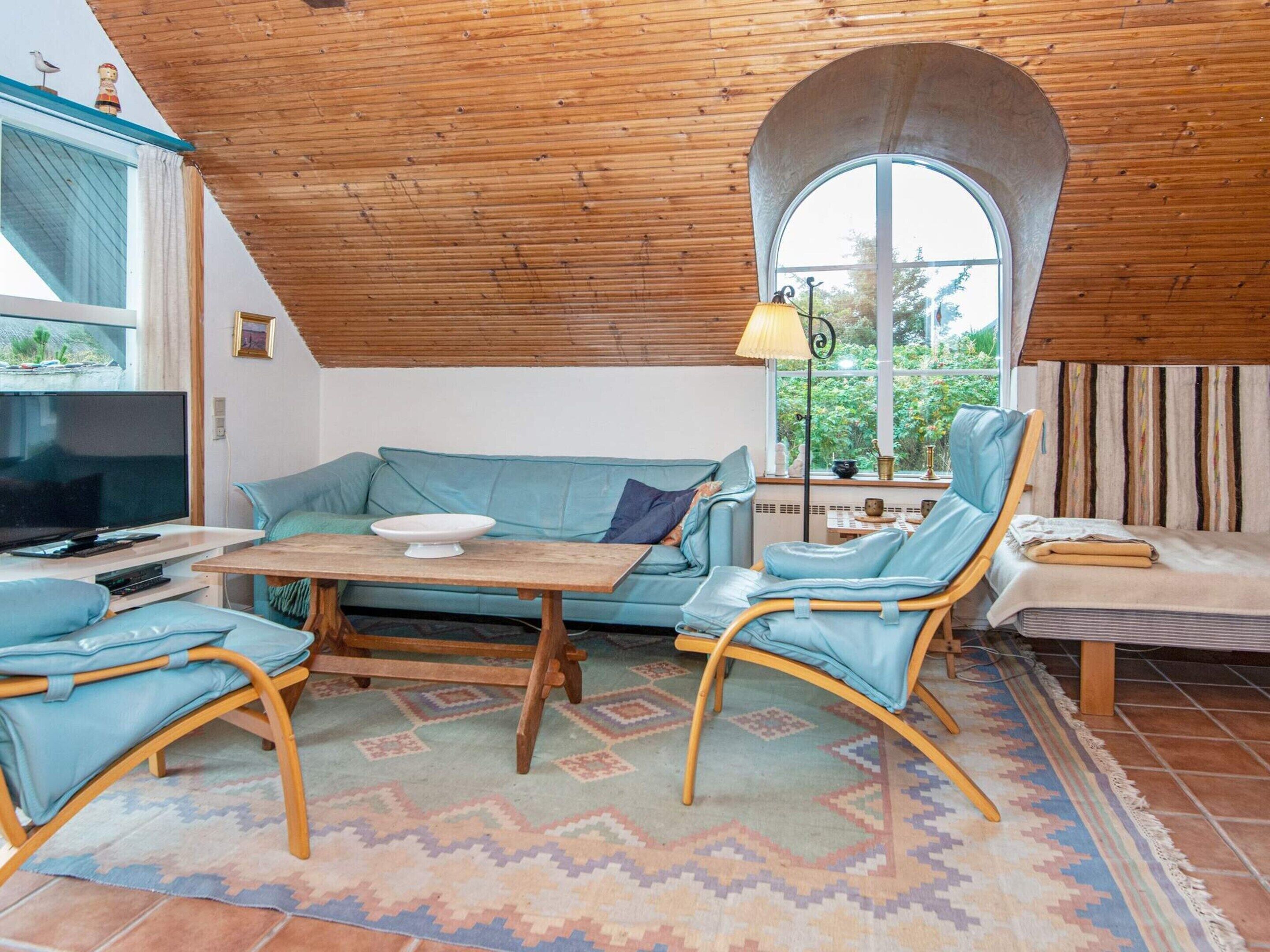 A cozy living room with a wooden ceiling, a large window, a television, a sofa, two chairs, and a table.