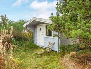 A small cabin with a white door and window, surrounded by dense greenery and a wooden bench.
