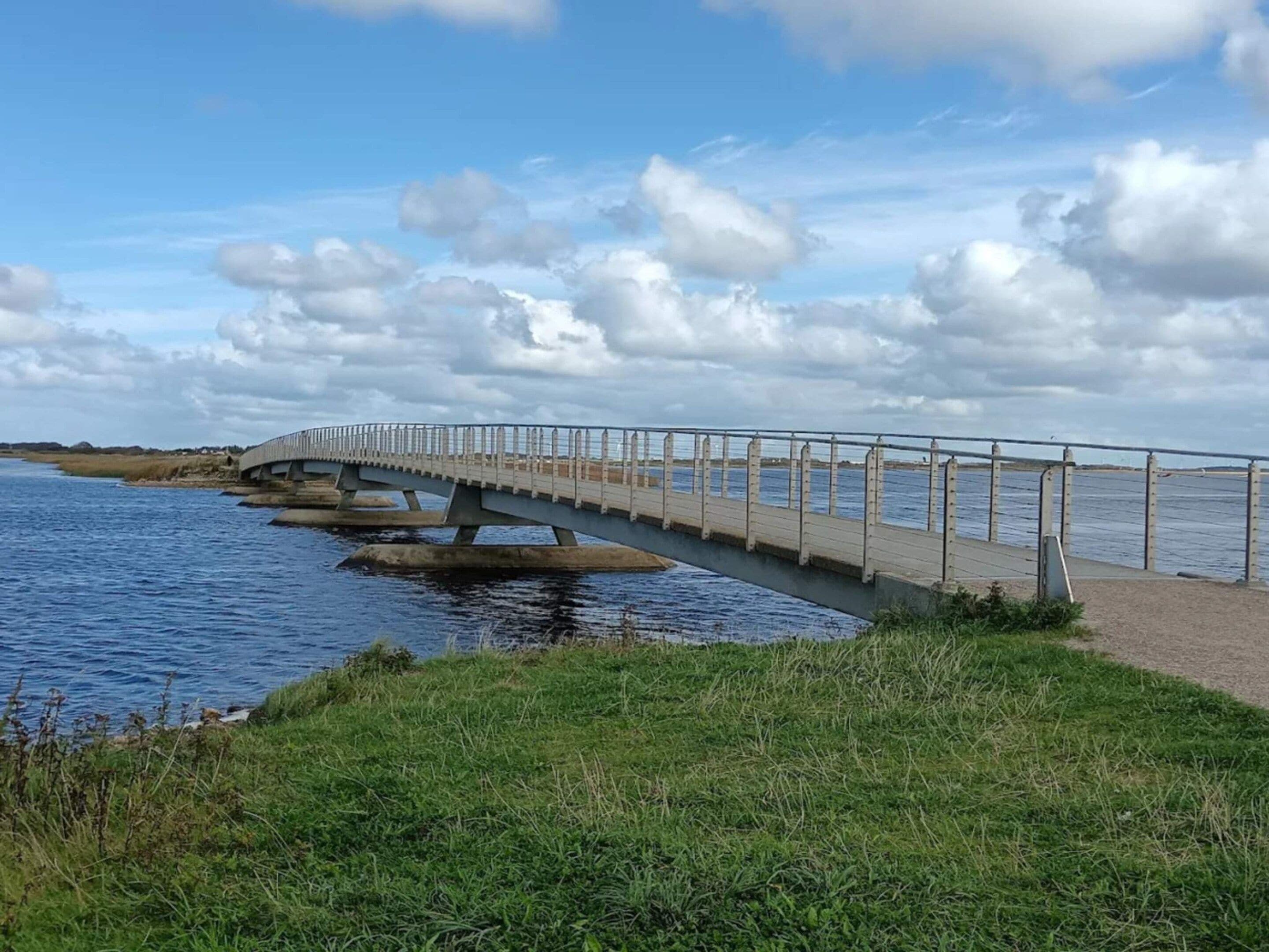 A pedestrian bridge over a body of water with a grassy bank on one side and a cloudy sky above.