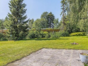 A well-maintained grassy area with a stone patio, surrounded by tall trees and a wooden pergola structure. - 4 Person Holiday Home in Vaeggerlose (Vaeggerlose)
