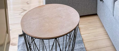 A round wooden table with a black wireframe base, positioned on a grey rug in a room with wooden flooring and a grey sofa.