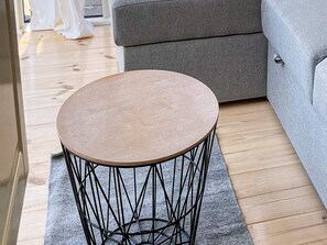 Una mesa redonda de madera con base de alambre negro, ubicada sobre una alfombra gris en una habitación con suelo de madera y un sofá gris.