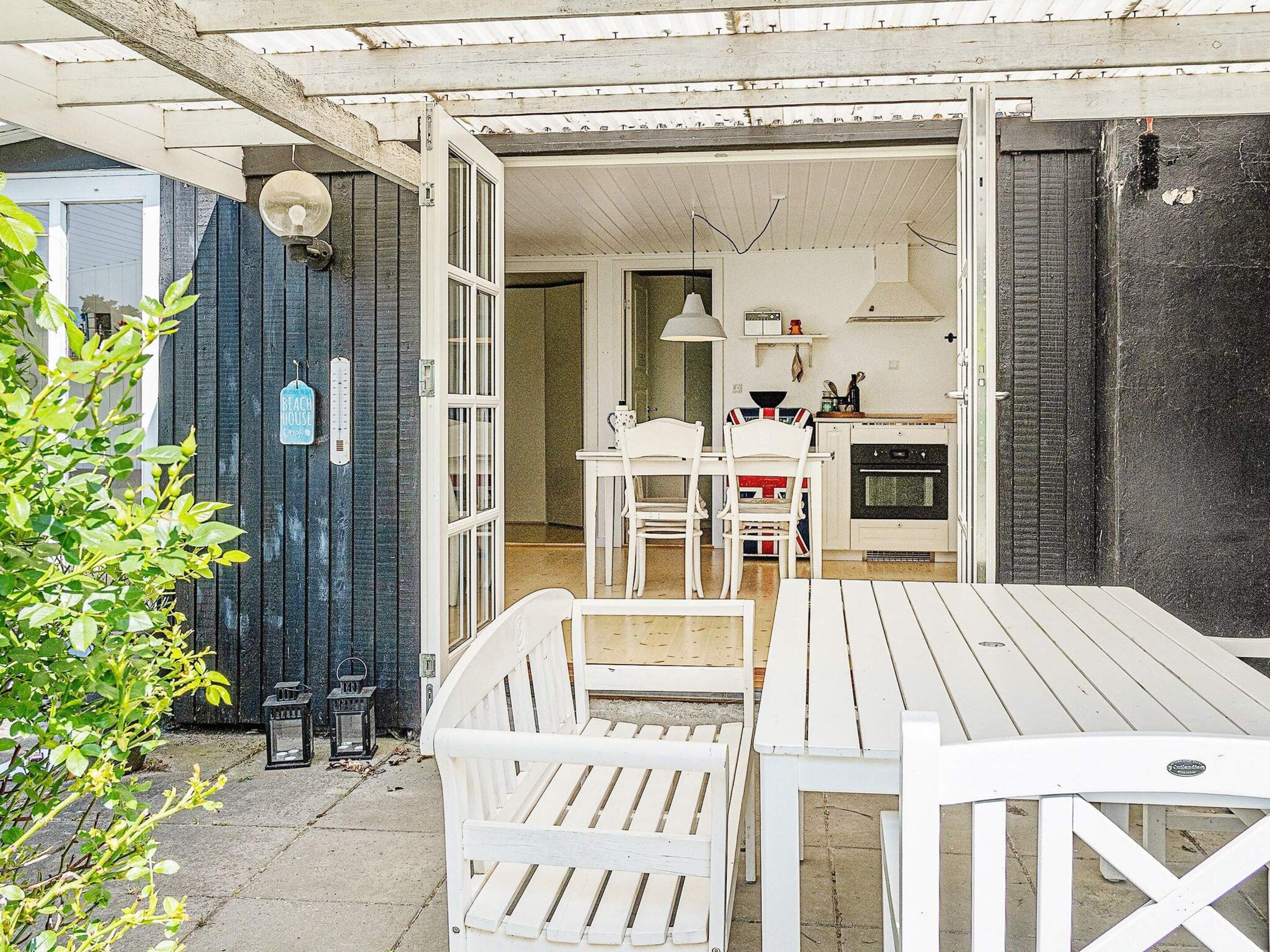 Une terrasse ouverte avec des meubles en bois peints en blanc, une vue sur une cuisine équipée d’une cuisinière et d’une table à manger, et une porte bleu foncé ornée d’un thermomètre.