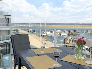 A table with a glass of red wine and a vase of flowers, overlooking a marina with boats and a beach.