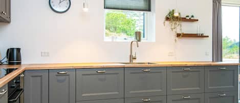 A modern kitchen with grey cabinets, a wooden countertop, a built-in sink, a clock, and a window with blinds.