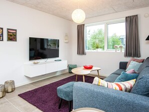 A modern living room with a flat-screen TV on a white cabinet, a grey sofa with colorful cushions, a small round table with a red bowl, and a purple rug.