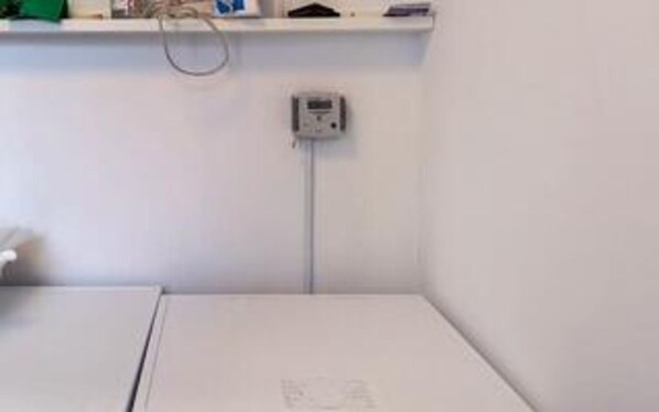 A room with a white bed, a small table, a wall-mounted electrical outlet, and a shelf with items. - 5 Star Holiday Home in Romo (Rømø)