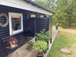 A wooden house with a black exterior, a window with a white frame, and a door. There are potted plants on the porch and a blue pot with red flowers in the background. - 6 Person Holiday Home in Hals (Hals)