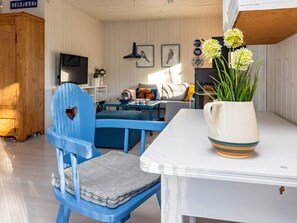 Una habitación con una silla azul, una mesa blanca y un jarrón con flores. Hay un sofá y una televisión al fondo.