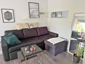 A modern living room with a dark grey sofa, a small side table, and a coffee table. There are decorative pillows, a framed picture, and a potted plant.