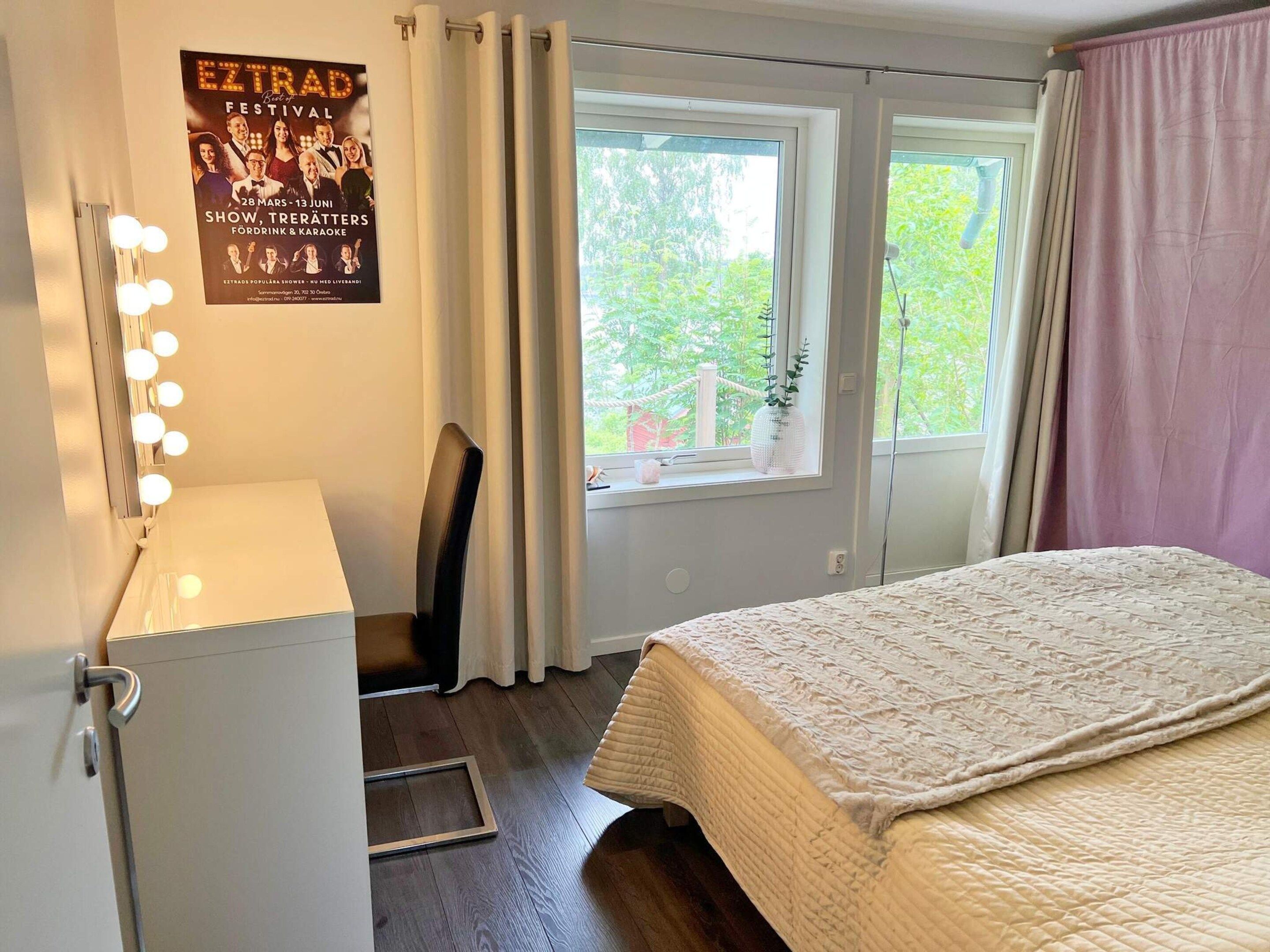 Une chambre d’hôtel avec un lit, un bureau doté d’une lampe, une affiche au mur et une fenêtre avec des rideaux.