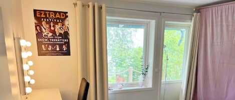 A hotel room with a bed, a desk with a lamp, a poster on the wall, and a window with curtains.