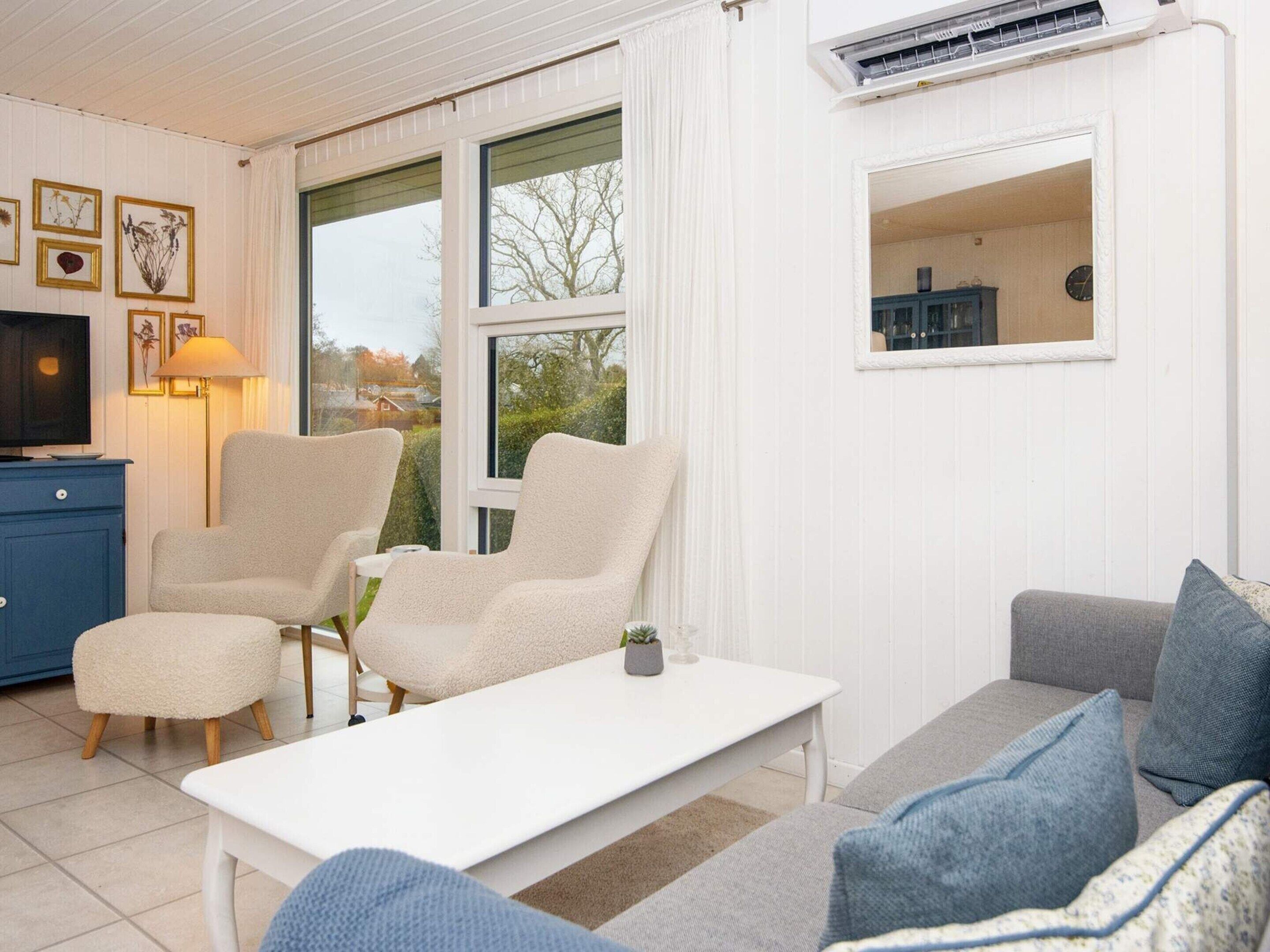 Un salon avec un canapé blanc, deux fauteuils, une table basse et un téléviseur posé sur un meuble bleu. Il y a une fenêtre donnant sur des arbres et une horloge au mur.