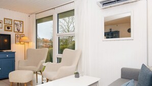 A living room with a white sofa, two armchairs, a coffee table, and a TV on a blue cabinet. There is a window with a view of trees and a clock on the wall. - 4 Person Holiday Home in Hejls (Hejls)
