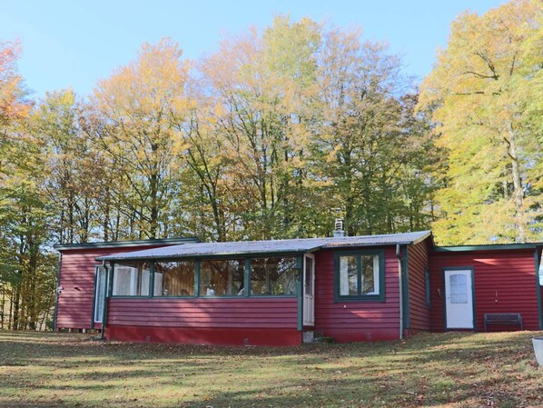 Une cabane rouge avec un petit porche et une cheminée, entourée d’arbres aux feuilles d’automne.