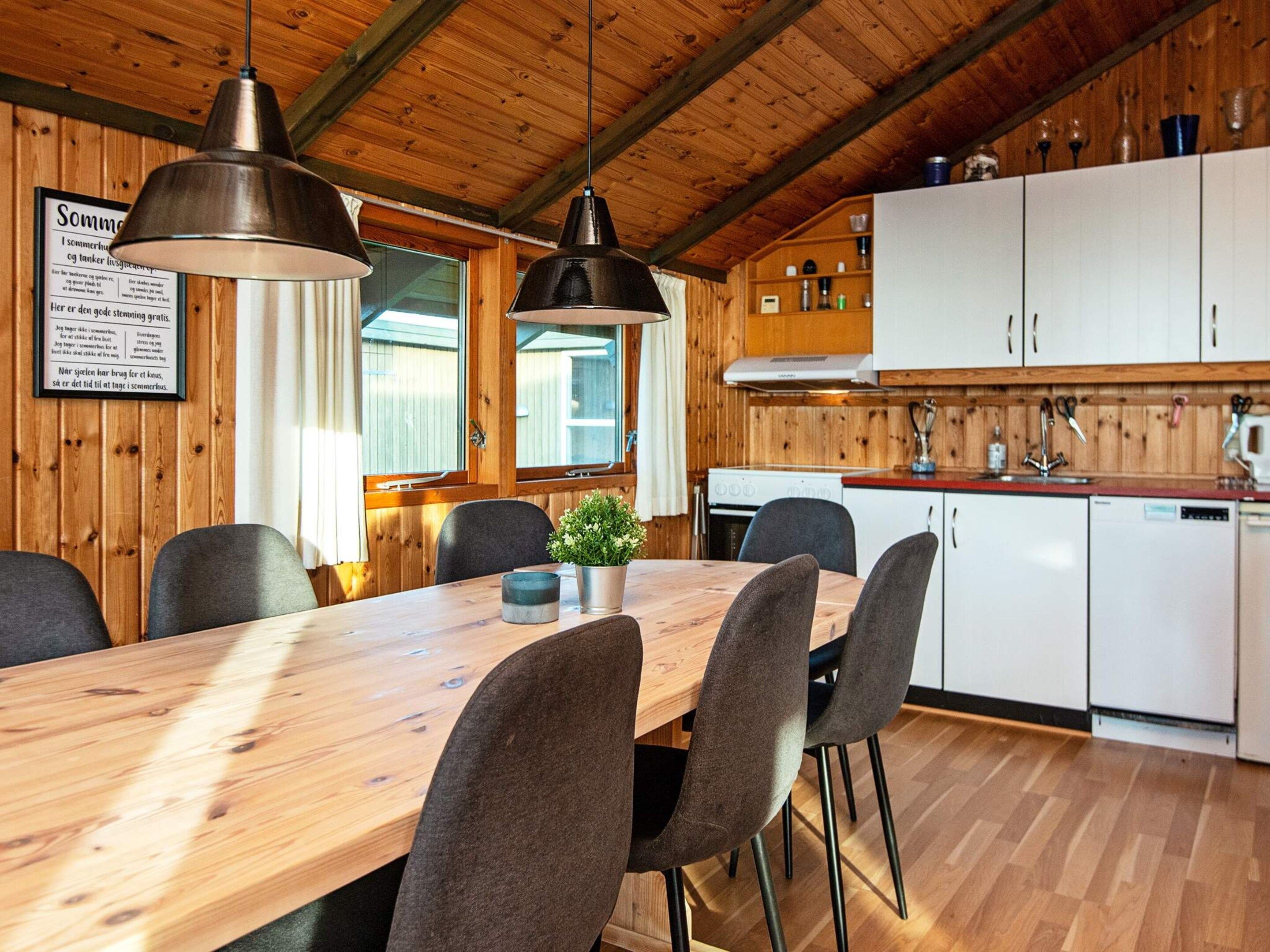 Un intérieur en bois comprenant une table à manger, des chaises, des luminaires suspendus, un coin cuisine avec des placards et un menu encadré accroché au mur.