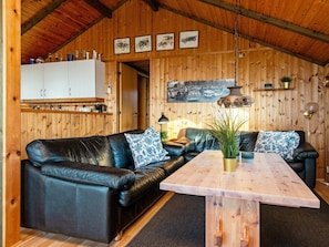 A cozy room with a black leather sofa, a wooden dining table, and a kitchen area with cabinets and a hanging light fixture. - 6 Person Holiday Home in Knebel (Knebel)