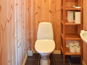 A small, wooden-paneled bathroom with a toilet, shelves with toilet paper, and a sink.