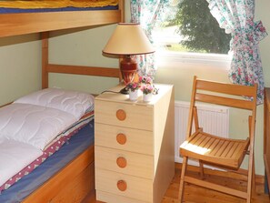 Une chambre avec un lit superposé, une commode en bois avec des tiroirs, une chaise, une lampe et une fenêtre avec des rideaux.