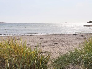 Une plage avec du sable et de l’herbe au premier plan, une mer calme et un bateau au loin.