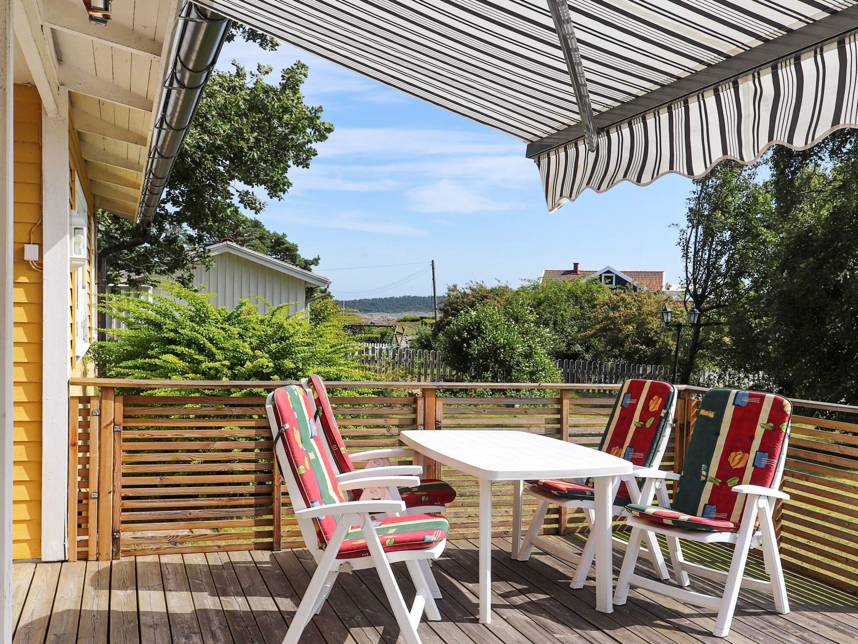 A wooden deck with a white table and two chairs, a covered area, and a view of a white building and greenery.