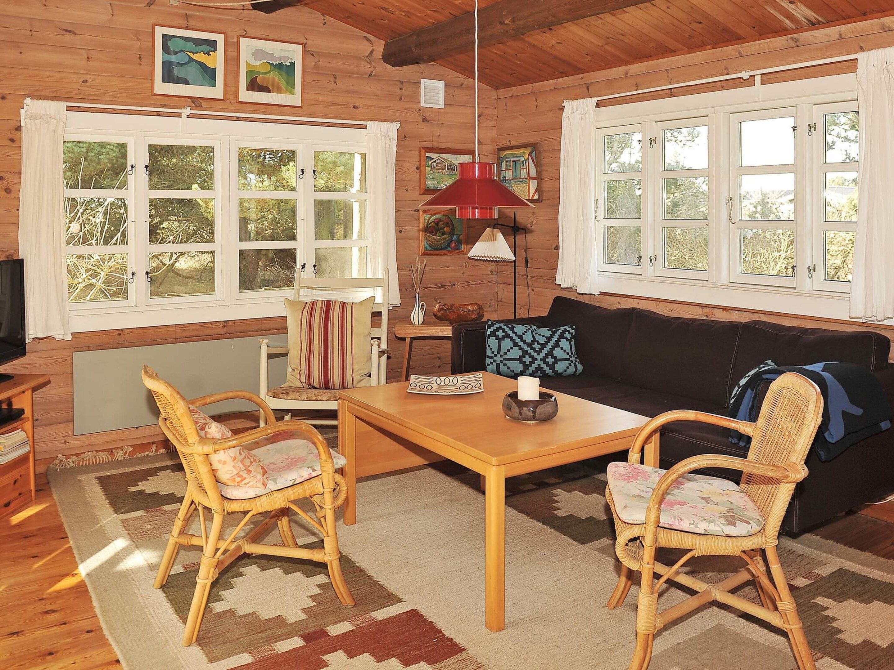 A cozy cabin interior with a wooden interior, a dining table, two wicker chairs, a sofa, and a red lamp.