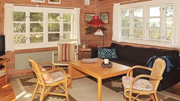 A cozy cabin interior with a wooden floor, a dining table, two wicker chairs, a sofa, and a television.