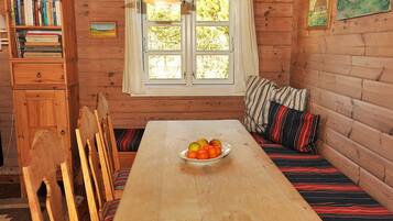 Interior de una cabaña de madera con mesa de comedor, sillas, un banco con cojines, estantería y ventana con cortinas.