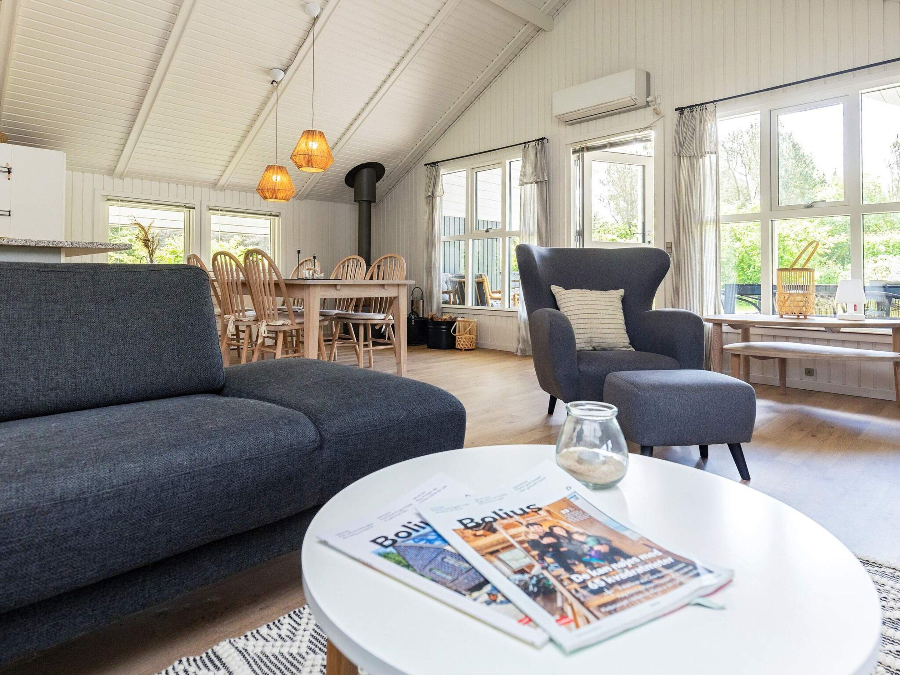 Una sala moderna con un sofá, un sillón y una mesa redonda con una revista. Hay una zona de comedor con sillas de madera y una mesa, y ventanas grandes con cortinas.