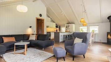 Un salón espacioso con un sofá de color gris oscuro, una mesita de centro redonda y un sillón gris. Se ve una cocina al fondo con una barra.