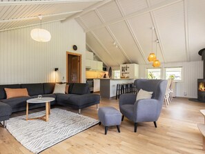 Un salón espacioso con un sofá de color gris oscuro, una mesita de centro redonda y un sillón gris. Se ve una cocina al fondo con una barra.