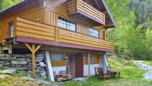 A two-story wooden cabin with a stone foundation, a balcony, and a small outdoor seating area.
