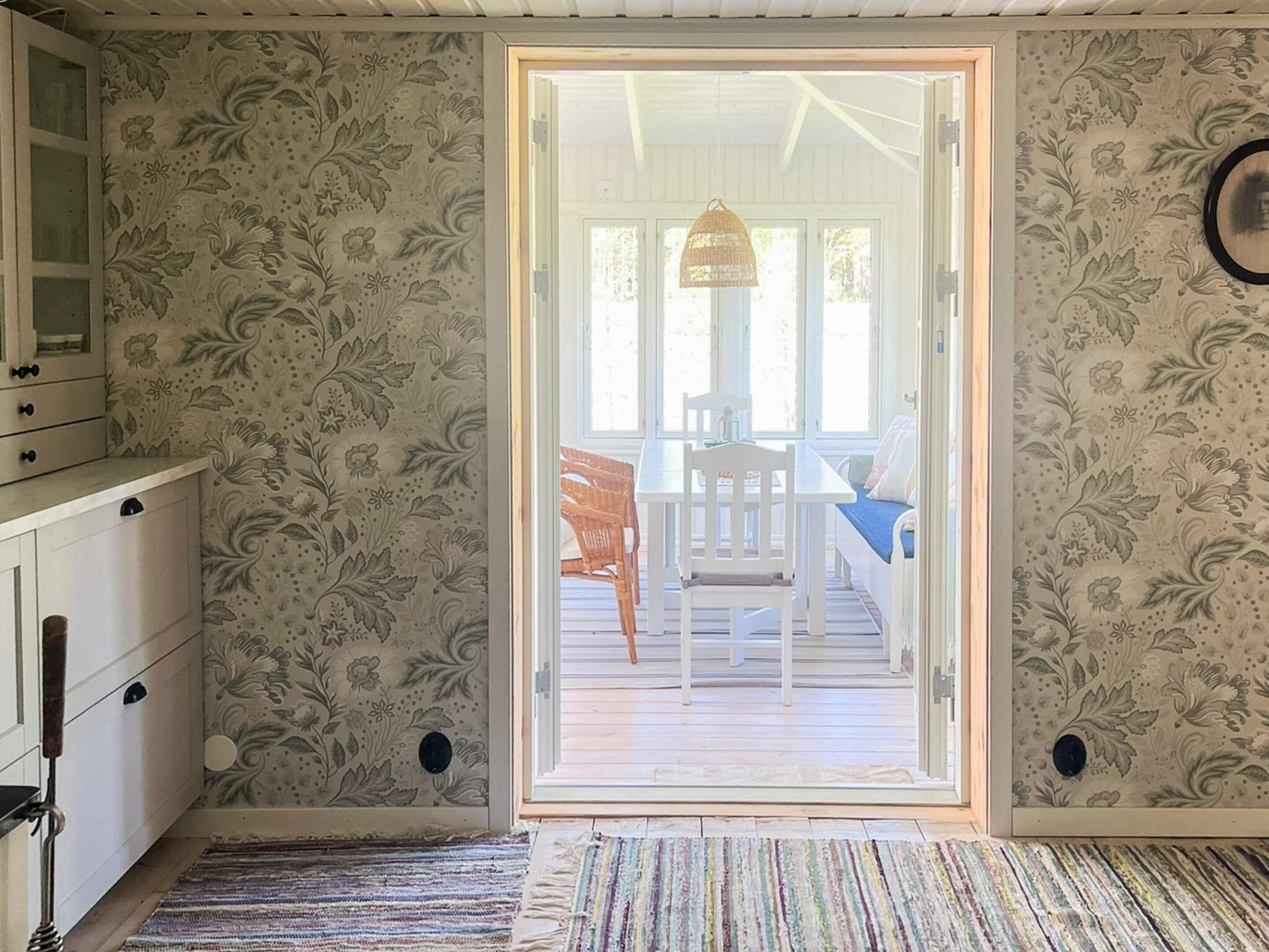 A room with a patterned wallpaper, a wooden floor, a white door leading to an outdoor area with a table and chairs, and a hanging light fixture.