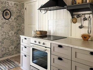 A kitchen with a stove, oven, and a black range hood. There is a clock on the wall and a set of hanging pots and pans.