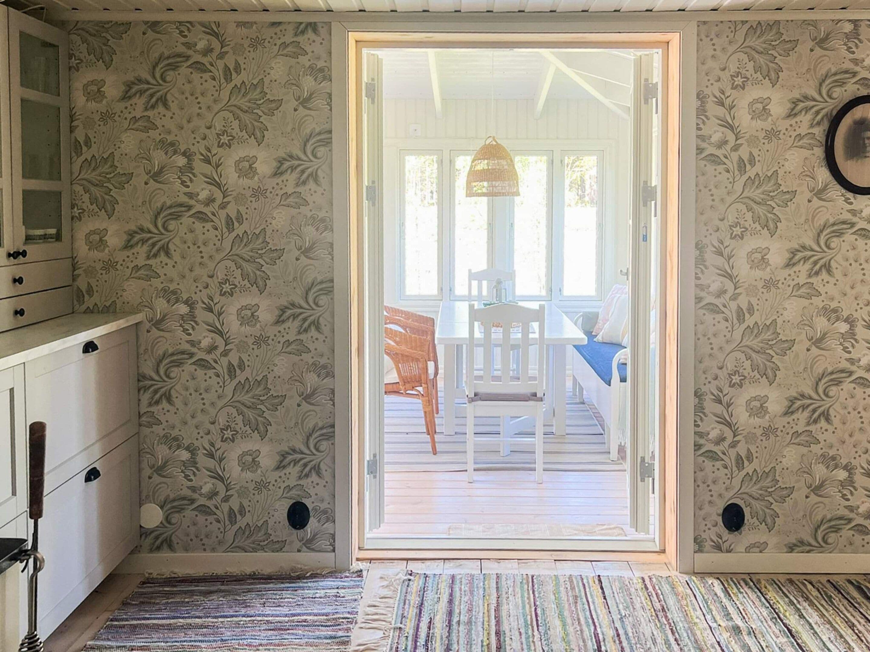 Una habitación con papel tapiz estampado, piso de madera, una puerta blanca que da a un comedor y un mueble bajo con cajones.