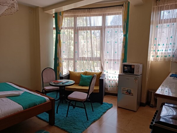 A compact room with a bed, a small table with chairs, a sofa, a microwave, and a window with curtains. - Cool Luxurious Studios (Nairobi)