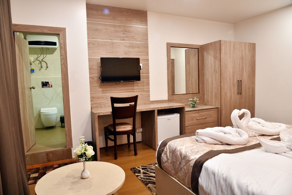 A hotel room with a bed, a desk, a chair, a television, a mirror, and a shower.