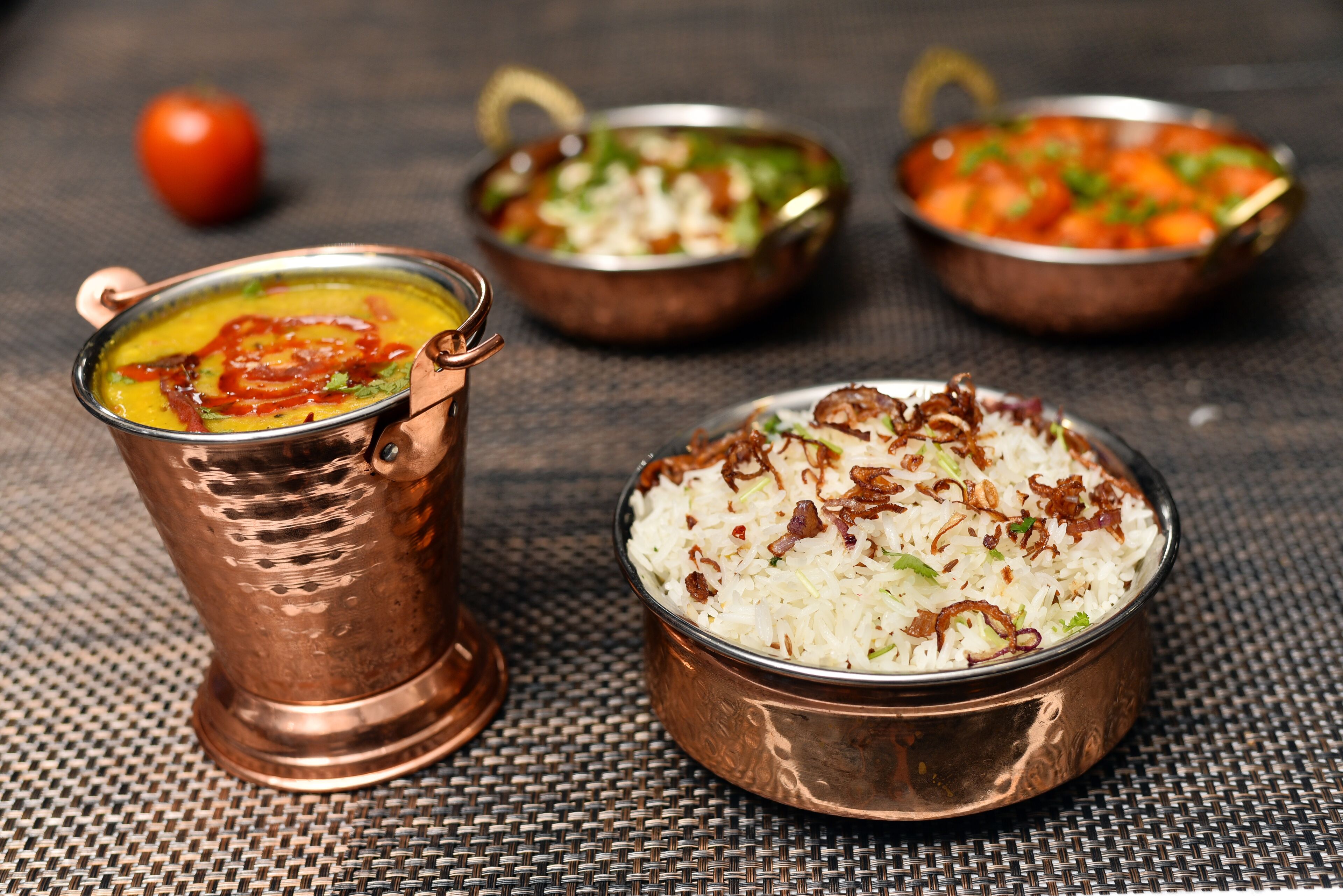 A table with three copper bowls containing different dishes, a glass of a yellowish beverage, and a tomato in the background.