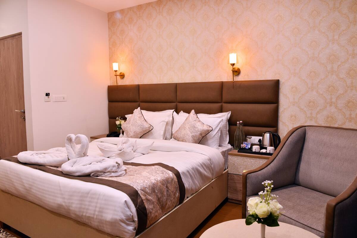 A well-appointed hotel room with a large bed, two bedside lamps, a nightstand with a coffee maker, and a seating area with a chair and a vase of flowers.