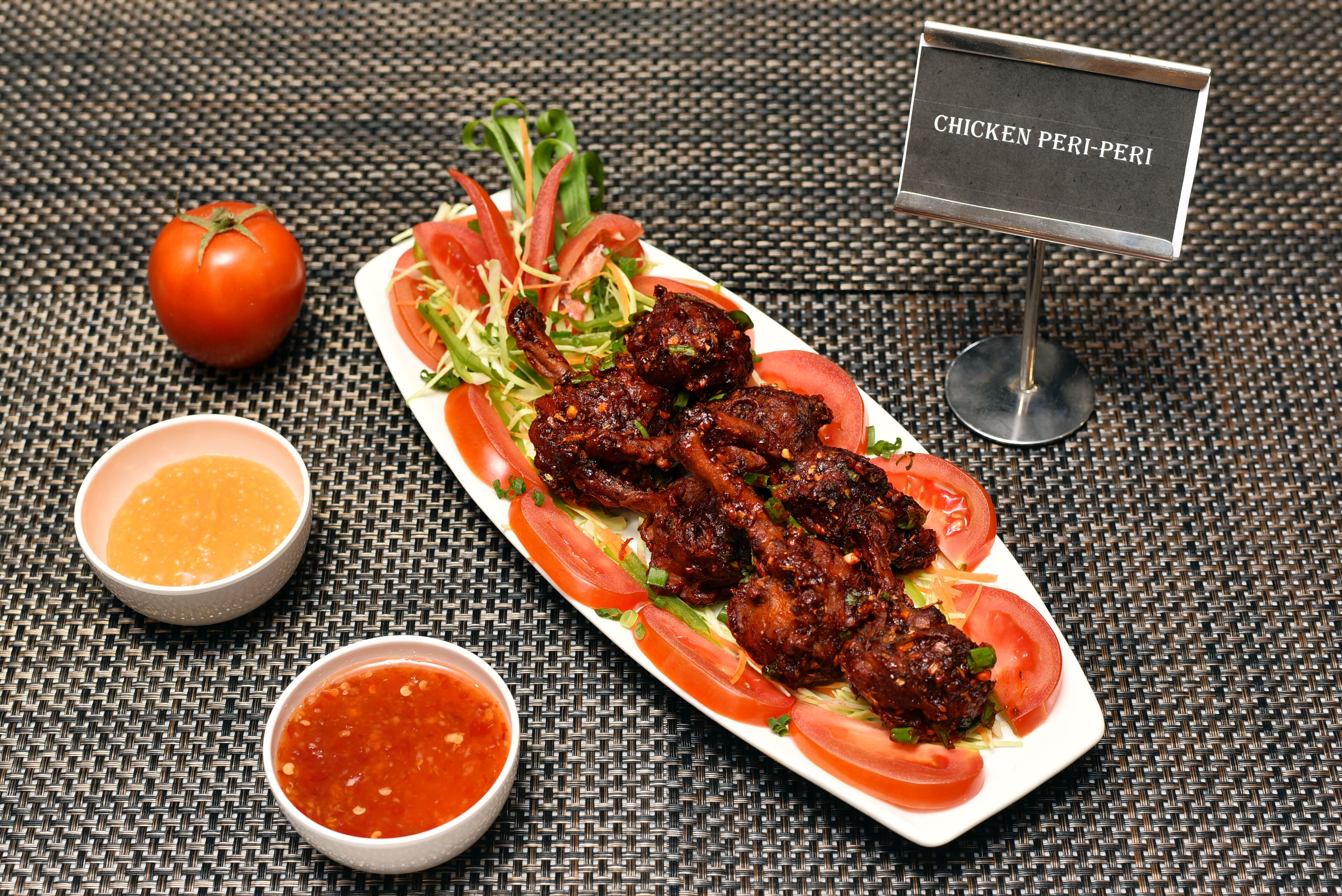 A plate of chicken peri-peri with a side of tomato and two dipping sauces.