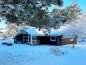 A snow-covered cabin with a garage, a fenced yard, and a small bush in the foreground.
