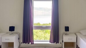 A room with two beds, a window with purple curtains, and a view of greenery and mountains.