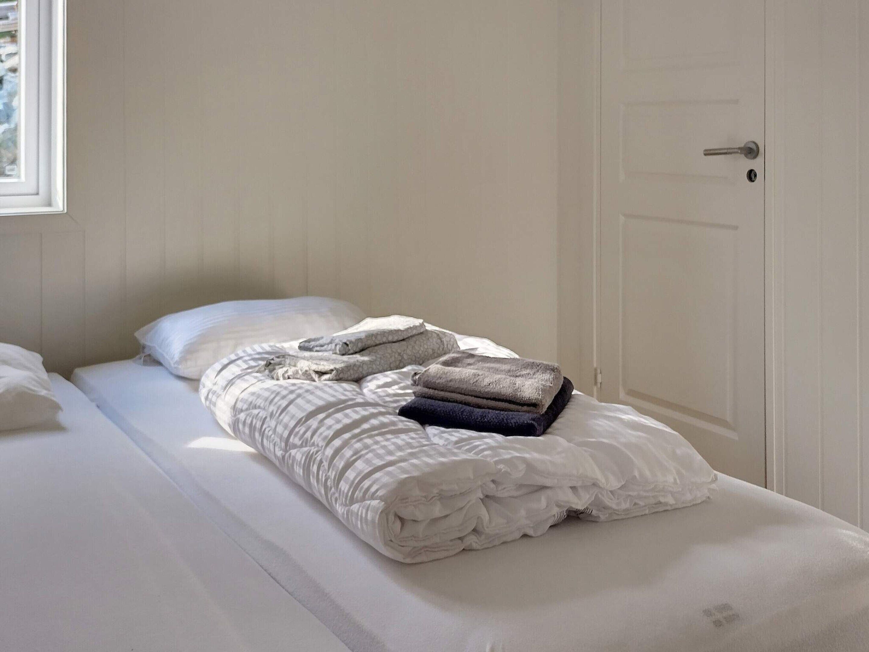 Una cama individual con ropa blanca de cama, una manta doblada y una toalla enrollada.