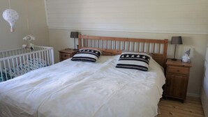 Een slaapkamer met een houten bed, wit beddengoed, twee nachtkastjes met lampen, een ledikant met een mobiel en een wandradiator.