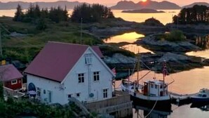 Un bâtiment blanc avec un toit rouge, un quai où sont amarrés des bateaux et une étendue d'eau calme, avec des montagnes en arrière-plan.