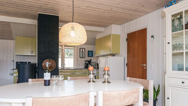 A dining area with a round table, chairs, a hanging light fixture, and a kitchen in the background.