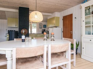 A dining area with a round table, chairs, a hanging light fixture, and a kitchen in the background.