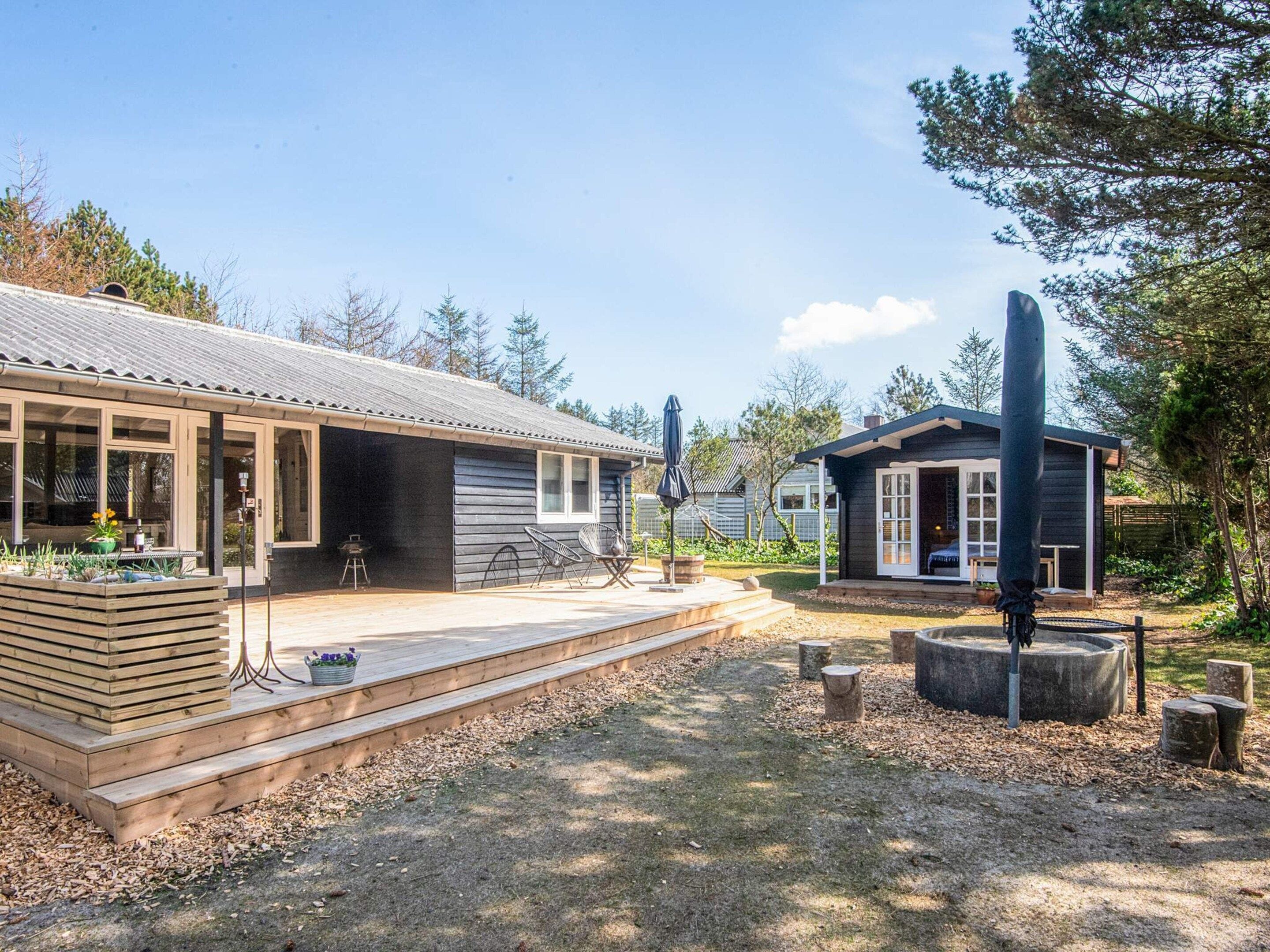 Una terraza de madera con mobiliario exterior, un pequeño jardín y un edificio con fachada oscura y techo inclinado.